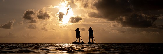 Bild zweier Menschen auf einem Brett stehend mit Paddeln auf dem Wasser vor dunkler Sonne
