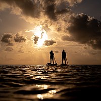 Bild zweier Menschen auf einem Brett stehend mit Paddeln auf dem Wasser vor dunkler Sonne