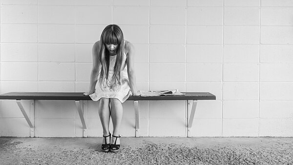 Schwarzweiß Aufnahme einer jungen Frau. Sie sitzt traurig und alleine auf einer Bank.